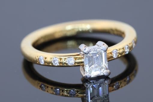 An elegant diamond engagement ring combining the timeless appeal of an emerald-cut diamond with the understated sophistication of 18kt gold. Its clean lines and subtle diamond detailing create a refined design that balances classic elegance with contemporary simplicity.