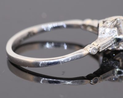 This Art Deco diamond engagement ring captures the elegance and geometric sophistication of the period. Combining a brilliant-cut centre diamond with baguette-cut shoulders and intricate engraved details, it is a distinctive jewel that celebrates the craftsmanship and design sensibility associated with Art Deco jewellery.