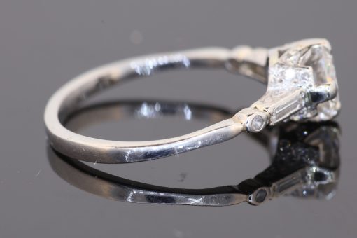 This Art Deco diamond engagement ring captures the elegance and geometric sophistication of the period. Combining a brilliant-cut centre diamond with baguette-cut shoulders and intricate engraved details, it is a distinctive jewel that celebrates the craftsmanship and design sensibility associated with Art Deco jewellery.