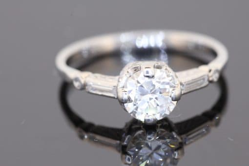 This Art Deco diamond engagement ring captures the elegance and geometric sophistication of the period. Combining a brilliant-cut centre diamond with baguette-cut shoulders and intricate engraved details, it is a distinctive jewel that celebrates the craftsmanship and design sensibility associated with Art Deco jewellery.
