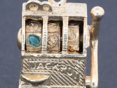 A wonderfully whimsical gold novelty charm in the form of a one-armed bandit, capturing the playful spirit of traditional gambling machines in miniature. The charm is particularly appealing for its working mechanism, transforming an instantly recognisable slot machine into an ingenious piece of novelty jewellery.