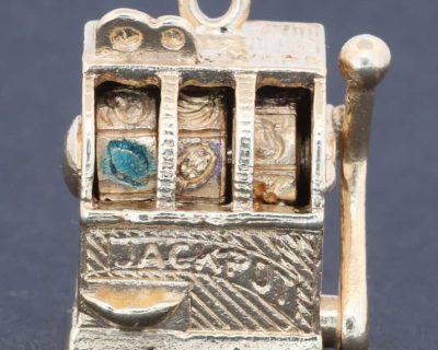 A wonderfully whimsical gold novelty charm in the form of a one-armed bandit, capturing the playful spirit of traditional gambling machines in miniature. The charm is particularly appealing for its working mechanism, transforming an instantly recognisable slot machine into an ingenious piece of novelty jewellery.