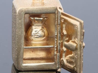 A charming and unusual gold novelty safe charm, designed as a miniature representation of a traditional safe and incorporating a delightful hidden surprise. Novelty charms of this kind transformed familiar objects into miniature works of jewellery, often with an element of humour or concealed detail. This example is particularly appealing for its clever association between a safe and its precious contents.